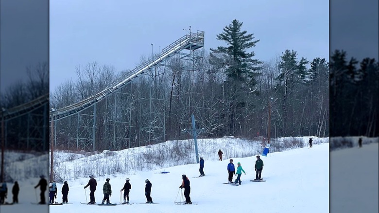 Skiers on a snowy slope with trees and a ski jump in the background
