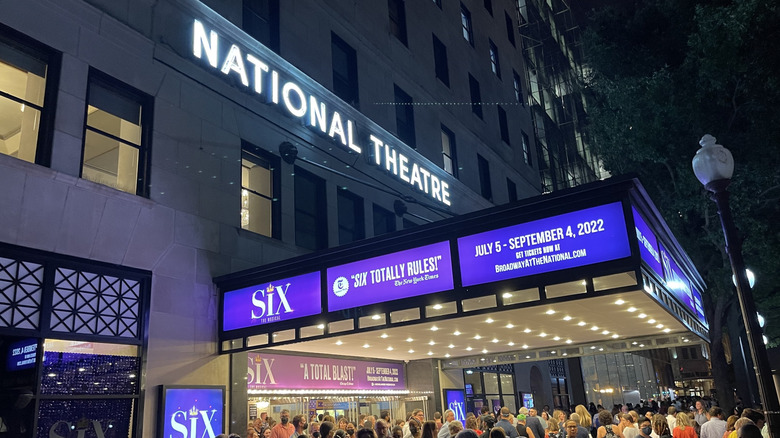 Exterior of the National Theatre at night, a white building with a marquee sign adevertising Six and a crowd milling beneath it