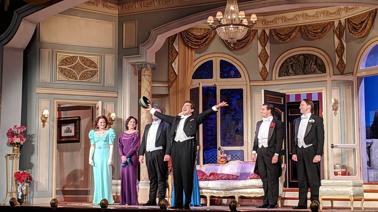 Performers on stage at Walnut Street Theatre, with four men in tuxes and women in blue and purple dresses in front of blue walls with grown accents under a chandelier