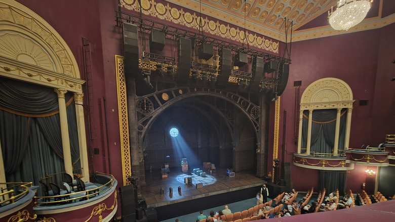 Stage of the National Theatre, surrounded by maroon walls with yellow and gold accents with a bank of speakers above the stage and people in seats at the bottom of the frame