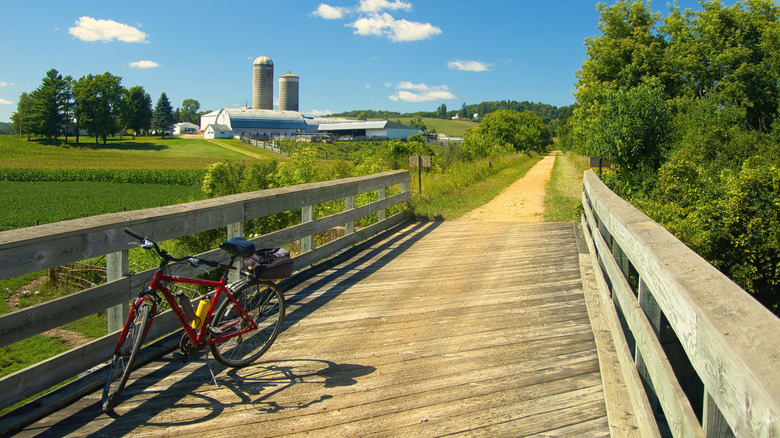 An empty red bike sitting along a trail over a bridge with farm buildings in the background