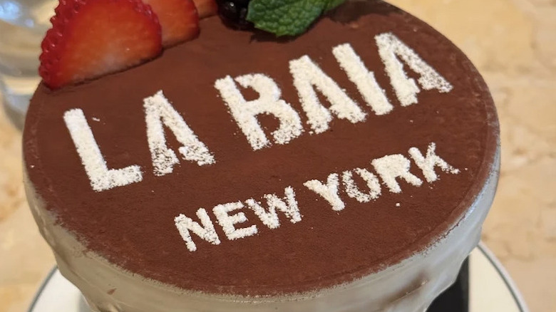Close up of Tiramisu dessert in a glass dish at Osteria La Baia with "La Baia New York" written on cocoa powder and strawberries visible at the top