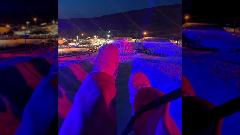 Nighttime view from a lift at Blue Mountain Resort, with legs in the foreground and colorful lights on the snow tubing lanes below