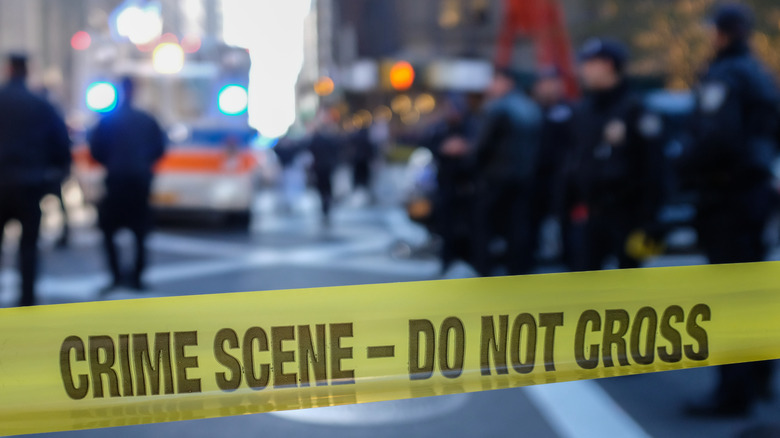 A city street blocked off by yellow crime scene tape with blurred out police officers in the background