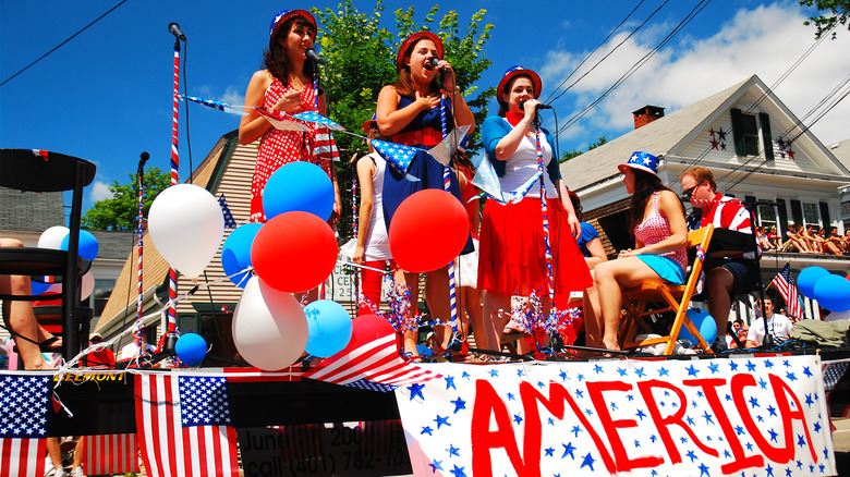 Three young women sing into microphones on a stage with red white and blue balloons and a sign that reads "America" in Bristol, Rhode Island