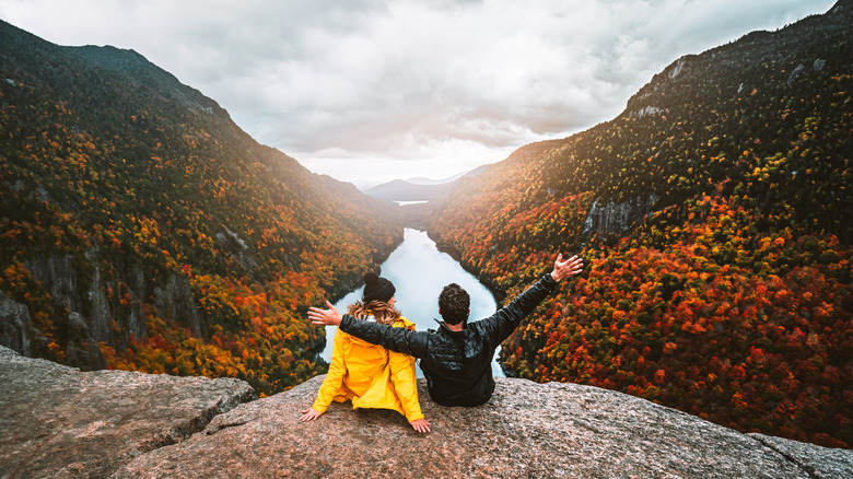 A man and a woman sit on a cliff overlooking a river flanked in fall foliage