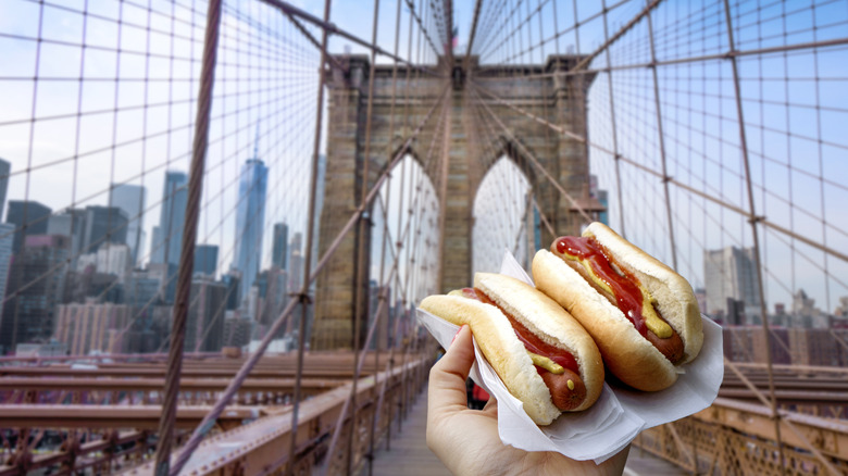 Hand holding two hot dogs with views of Brooklyn Bridge