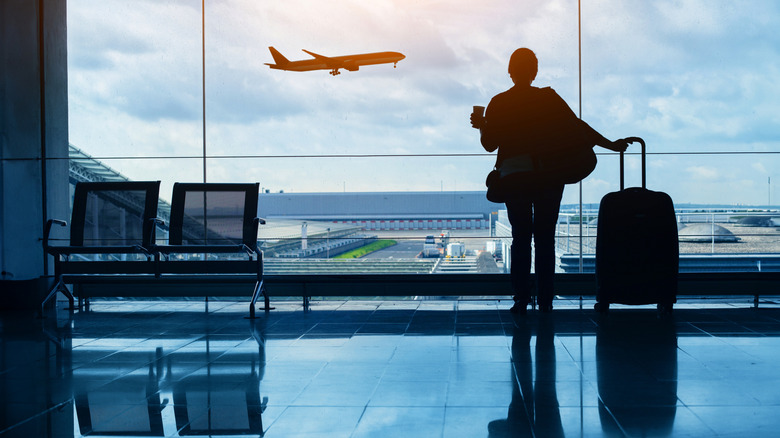 A woman holding luggage and a cup looks out the window at a plane taking off at an unidentified airport