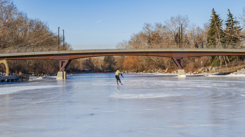 Man ice skating on frozen Canadian river with trees and bridge in background