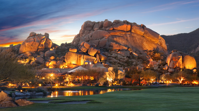 Nighttime exterior view of Boulders Resort & Spa Scottsdale, a five-star luxury resort located in the town of Carefree in central Arizona, with golf greens and a pond in the foreground and the red rock landscape in the background.