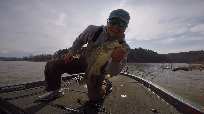 A smiling man in a ball cap and sunglasses kneeling on a boat holding a fish.