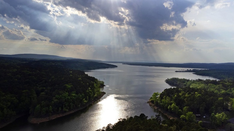 A view of Nimrod Lake stretching into the distance with hills and trees on either side and a cloudy sky with sunrays beaming down.
