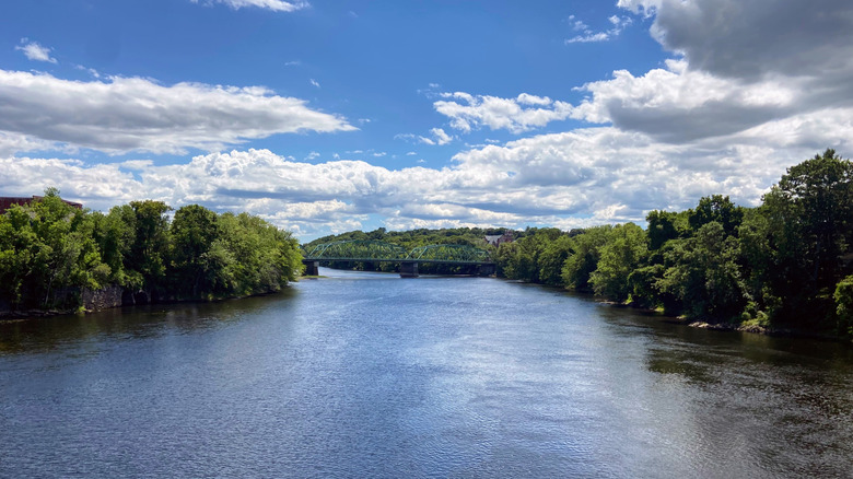 A bridge in the distance crossing over the Androscoggin River, which is lined with trees on a bright day in Auburn, Maine