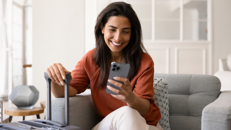 A smiling woman sits in an armchair holding her suitcase in one hand and her smartphone in the other hand, while making an online booking.