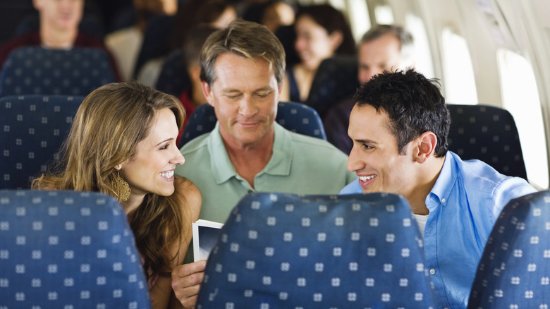 The interior of a commercial plane, where a man and woman talk over another man, who is stuck in between them in the middle seat and is reading a magazine.