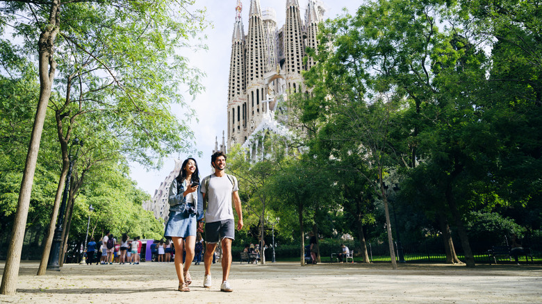 A couple walking in Barcelona between trees, with La Sagrada Familia in the background