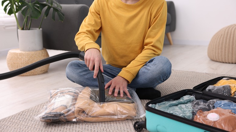 Male traveler sitting on the carpet vacuum sealing a bag of clothes next to a suitcase filled with clothes.