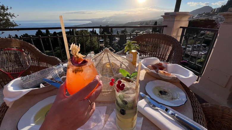 A hand holding up a cocktail at a table on the terrace of Bar Timeo in Taormina, Sicily, with a view of the sea in the distance