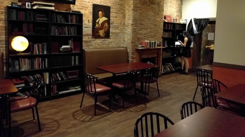 Interior of the Library on Carson, with a tan brick wall with a portrait of Shakespeare hanging beside a fireplace with books on its mantle between two black bookshelves, one with a person in a black dress standing at it, and red tables and chairs arranged on a wooden floor