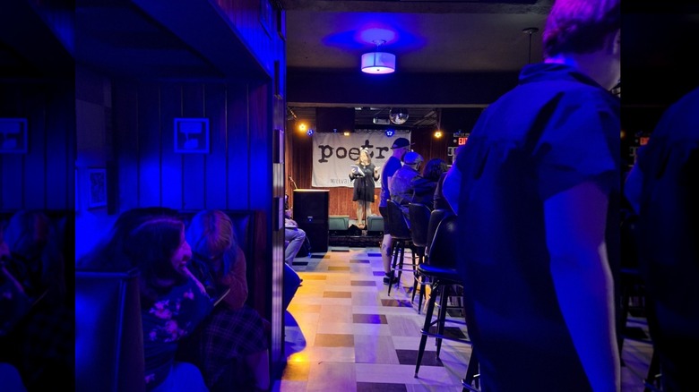Poet in a black dress reading on the stage at Poetry Lounge, with blue-lit patrons sitting at the bar along the right and in booths along the left and a black-and-white tiled floor