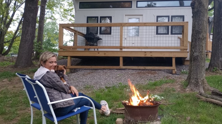 A smiling women holding a dog sits in front of a cabin in the trees.