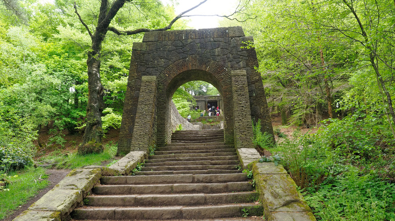 an old stone archway stands over a set of stairs in the middle of a leafy dense green forest; the stairs lead to a historic stone building at the top with people gathered there