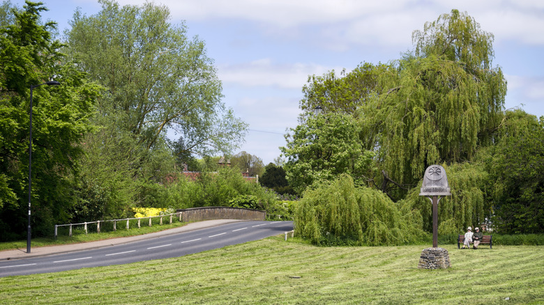A man and a woman sitting on a bench near a village sign and the bridge over the River Stour at Long Melford, Suffolk
