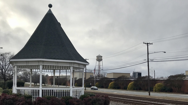 A white, pointy roofed gazebo near the railroad tracks in Landis, North Carolina; the sky is grey and there is a water tower in the background