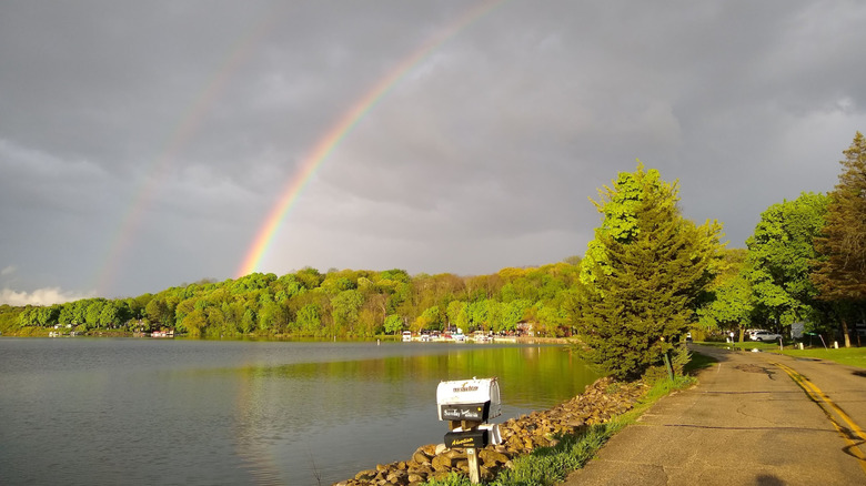 View of the waters, tree-lined shoreline, gray skies with a rainbow, and road around Whitewater Lake, Wisconsin