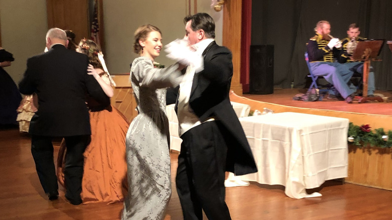 People dancing during the South Charleston New Years Eve Grand Ball, with a woman in a white dress dancing with a man in a tux in center frame, and a band in period costumes on the stage in the background