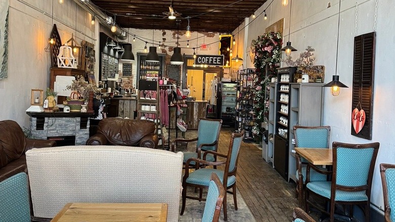 The Village Cup in South Charleston, a white-walled room with wood floors and ceilings, with shelves, wooden tables, and blue chairs along the walls and a cafe in the back with a sign reading "Coffee".