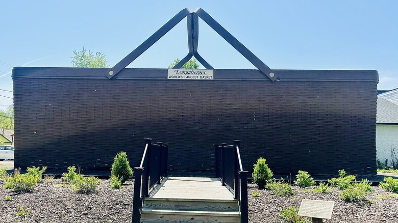 Large basket with a metal sign reading "Longaberger World's Largest Basket" and a walkway leading to it lined in black railings, surrounded by dirt with green shrubs, under a blue sky