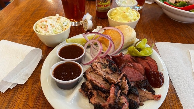 A plate of barbecue meat selections and sauce with a roll and side dishes of cole slaw and potato salad
