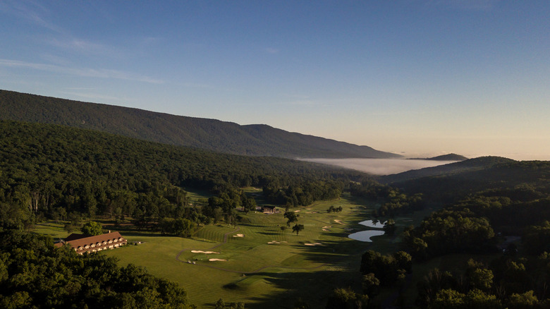 Aerial view of Cacapon State Park's golf course and lodge at sunrise, with mountains and blue sky in the background