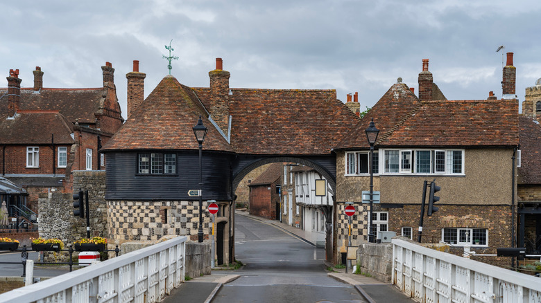 A bridge leads to a historical gate with a tiled roof beneath a cloudy sky in Sandwich, U.K.