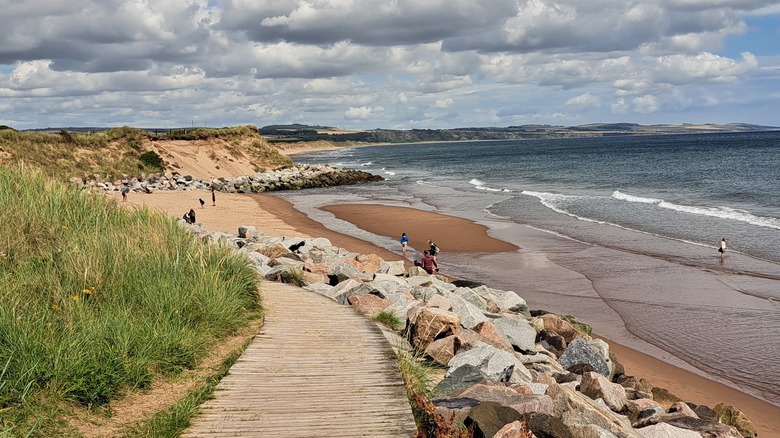 Boardwalk flanked by rock banks leading to sandy beach with tidal pools.
