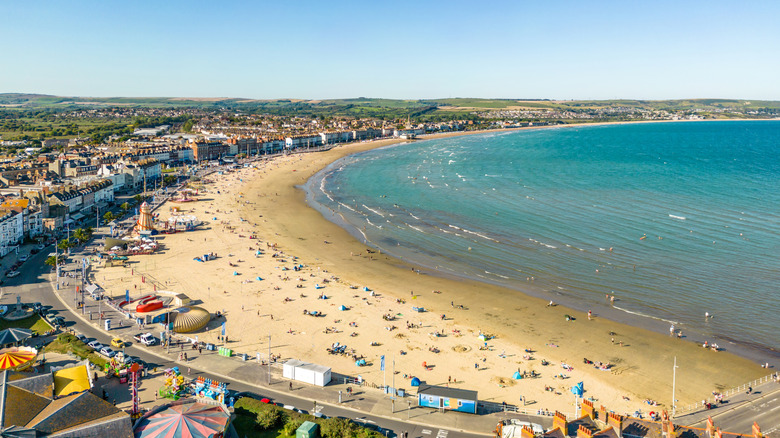 Buildings along the curved shoreline of Weymouth Beach with sunbathers and blue water under a blue sky