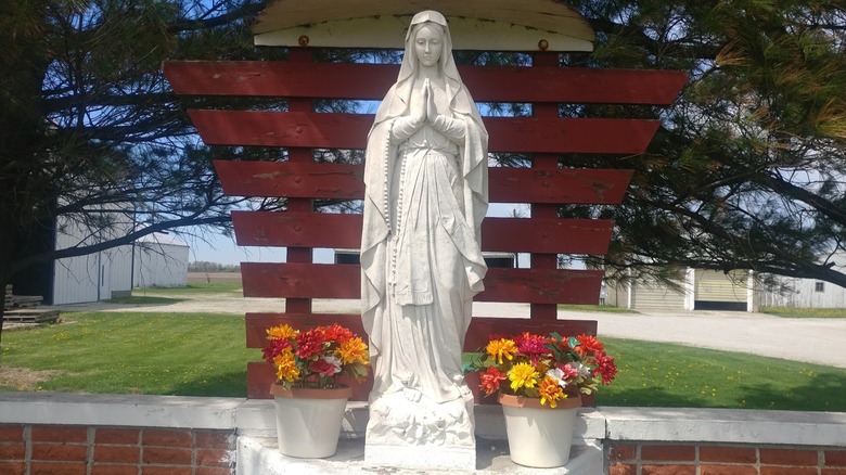 Marble statue of Virgin Mary praying in front of red wood and next to flower pots