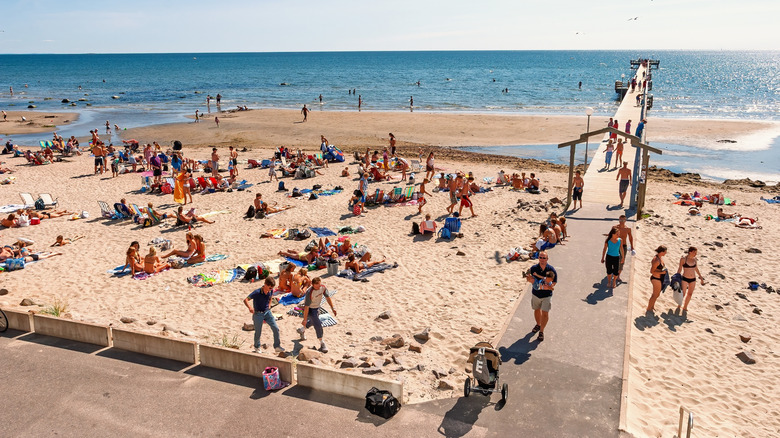 Lots of people laying and walking on the sand on a sunny day at Skea Strand with the thin pier going off into the sea