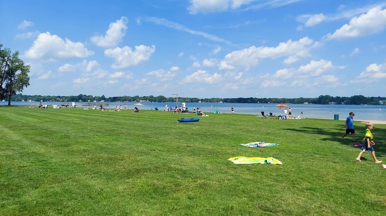 A crowded swim beach at Lake Lansing in Haslett, Michigan, featuring a large green space and a lake in the distance.