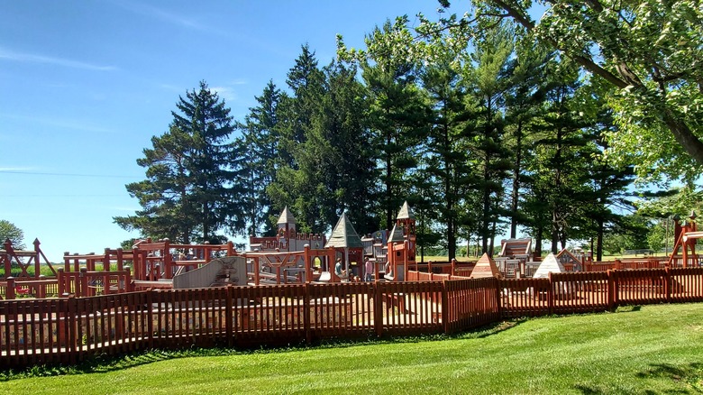 The large wooden playground at Woodland Park in Ithaca, with a low wooden fence around it and tall pines behind on a sunny summer day