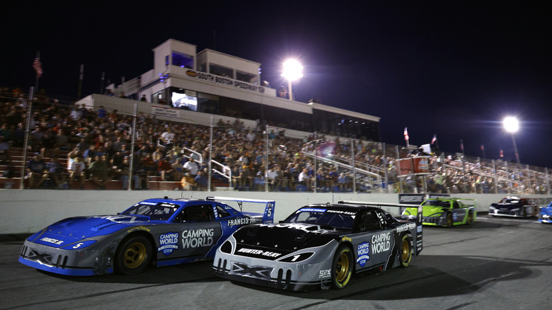 Two branded race cars, one blue and one gray, in the foreground of the track at South Boston Speedway with crowded bleachers.