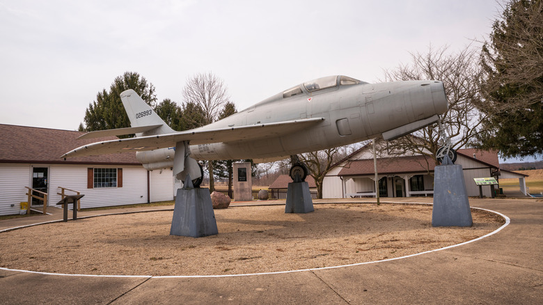 Silver single-person plane ongray blocks, with white-sided brown-roof buildings behind it and to the left and trees in the background, under a gray sky