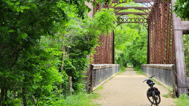 Bicycle rests on a historic rust-colored iron truss bridge surrounded by dense green woodland at the Ottawa trailhead of the Prairie Spirit Trail in Kansas