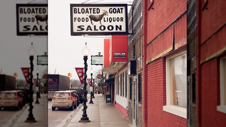 Sign of the Bloated Goat Saloon in Fowlerville over a sidewalk with cars parked beside it under a gray sky