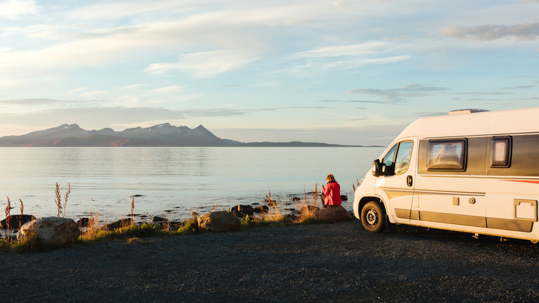 Person standing at the edge of a lake in front of a small RV with mountains on the other side.