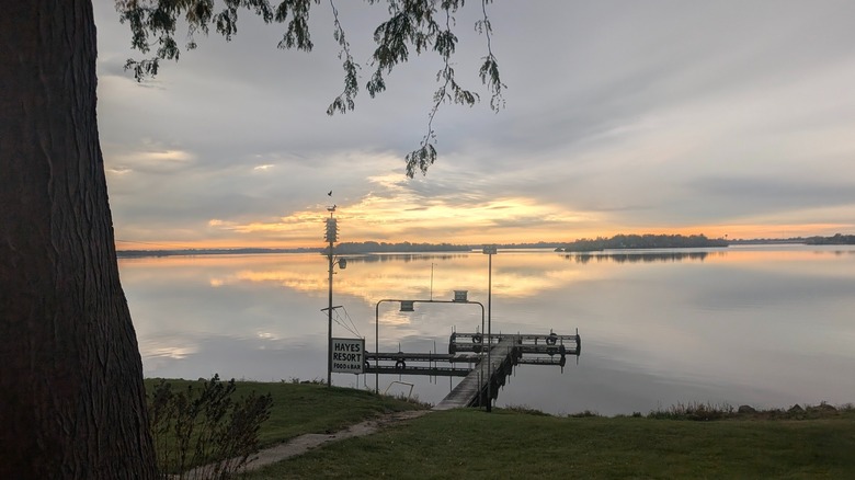 A quiet dock stretching from the grassy shore on Fox Lake with super calm waters reflecting the colorful evening sky