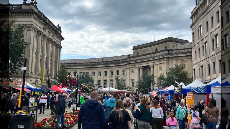 People walking past food stalls flanked by colonnaded stone buildings in Bolton