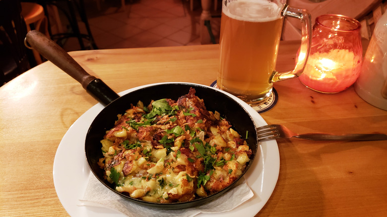 A hearty Austrian meal consisting of potatoes, bacon, cheese, and spatzle served in a pan with a cold beer