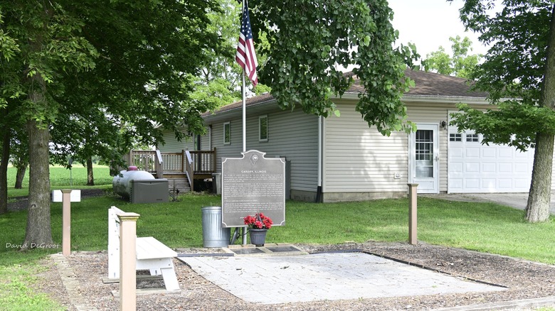 Historic marker for Cardiff, Illinois, with an American flag hanging over a plaque with a metal trash can, red flowers, and a white bench, in front of a white-sided house surrounded by trees